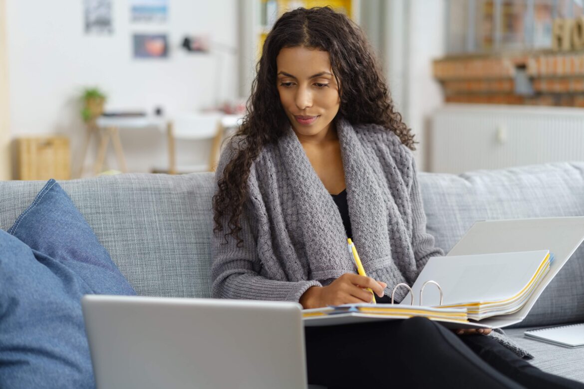 Woman sitting on couch with laptop and binder, working.