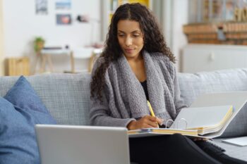 Woman sitting on couch with laptop and binder, working.